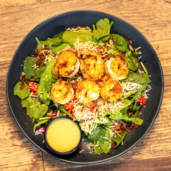 A black bowl of shrimp salad with spinach, cheese, bacon bits, and a side of dressing.