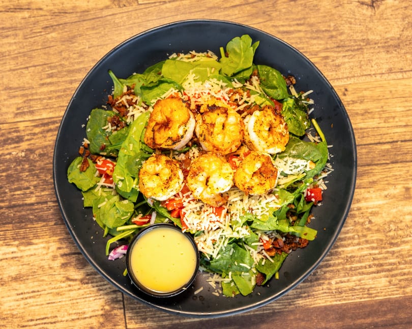 A black bowl of shrimp salad with spinach, cheese, bacon bits, and a side of dressing.