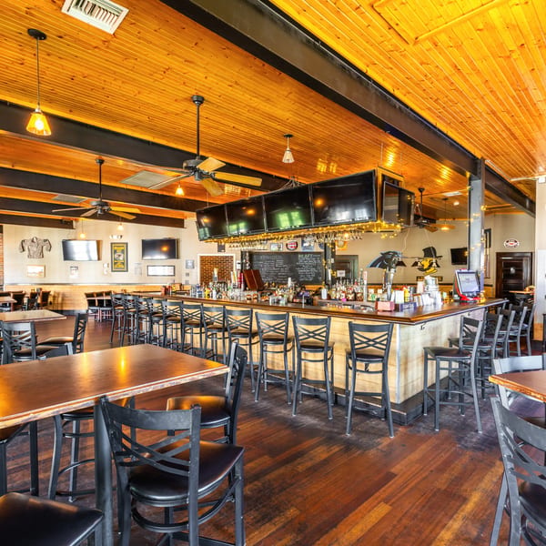 Spacious bar and dining area with wooden tables, black chairs, and a long bar with multiple TVs.