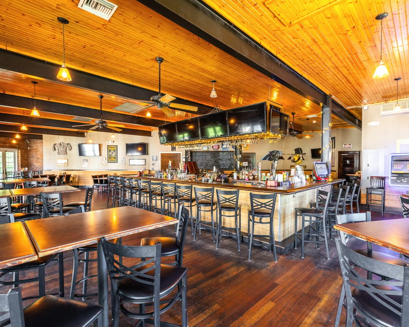 Spacious bar and dining area with wooden tables, black chairs, and a long bar with multiple TVs.