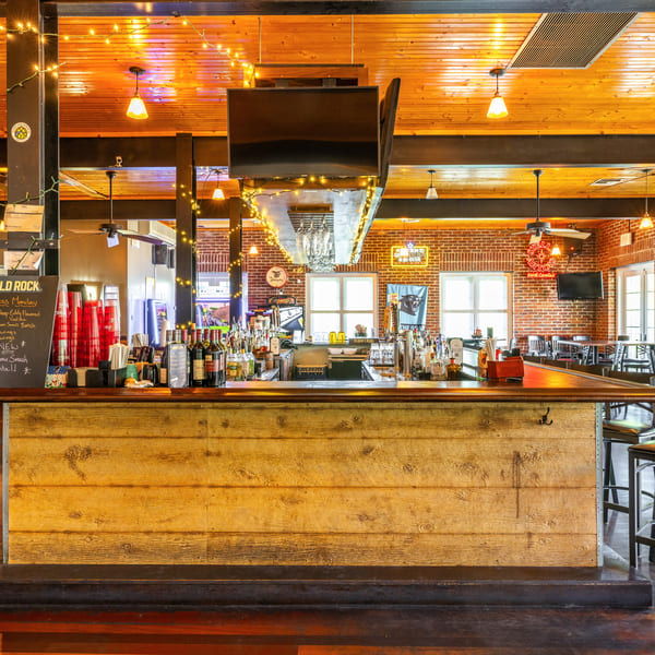A rustic wooden bar with bottles, glasses, and a menu board, in a restaurant with brick walls.