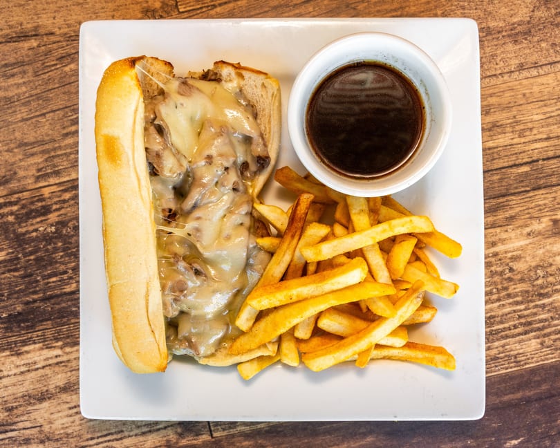 Cheesy Philly cheesesteak sandwich with a side of golden fries and a cup of au jus.