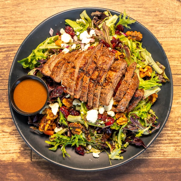 Grilled steak salad with mixed greens, feta, walnuts, cranberries, and a side of dressing.