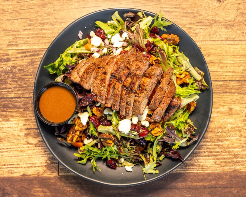 Grilled steak salad with mixed greens, feta, walnuts, cranberries, and a side of dressing.