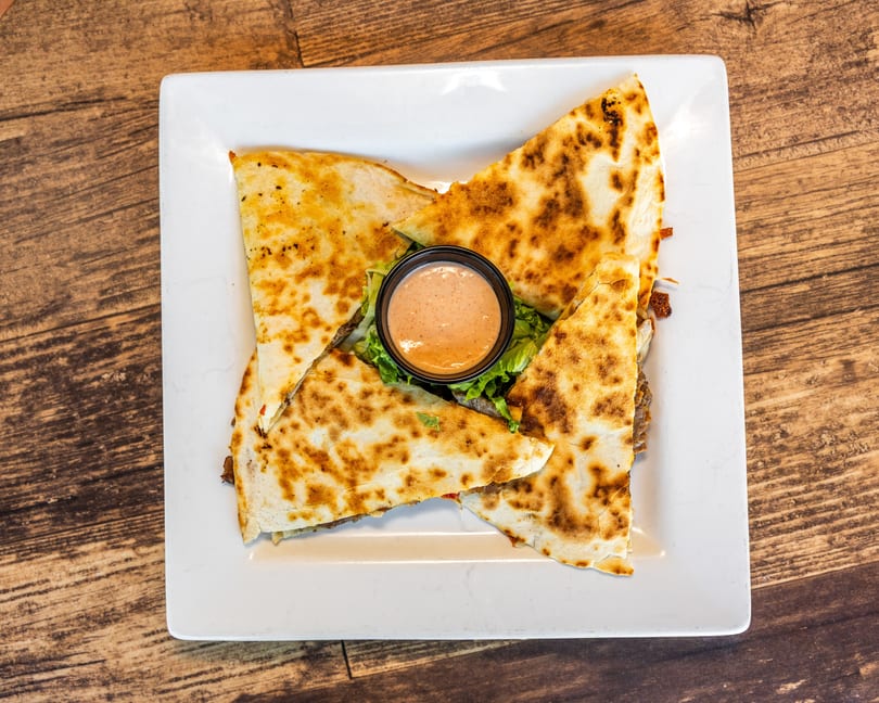 Four quesadilla wedges with a side of dipping sauce on a bed of lettuce, on a white plate.