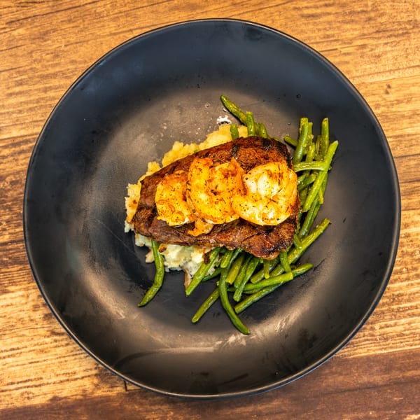 Steak and shrimp with mashed potatoes and green beans on a black plate.