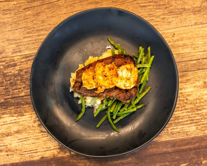 Steak and shrimp with mashed potatoes and green beans on a black plate.