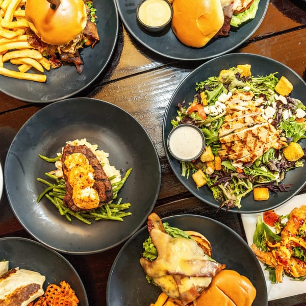 Overhead view of various dishes including burgers, salads, steak, shrimp pasta, and wraps on a dark wooden table.