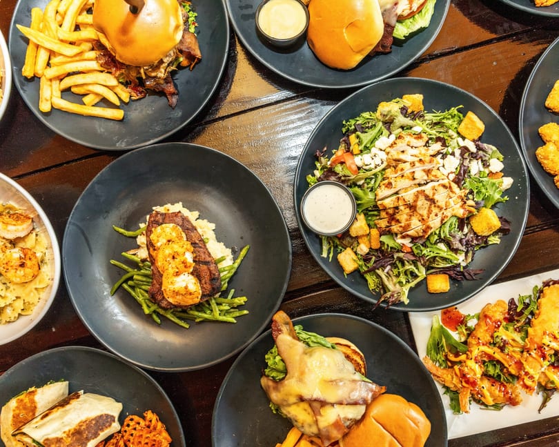 Overhead view of various dishes including burgers, salads, steak, shrimp pasta, and wraps on a dark wooden table.