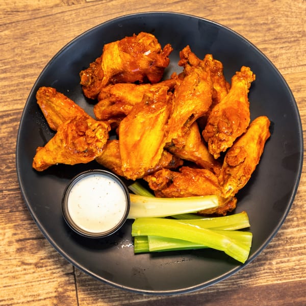 A black plate of buffalo chicken wings with ranch dressing and celery sticks on a wooden table.