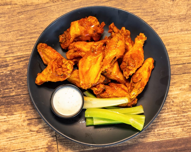 A black plate of buffalo chicken wings with ranch dressing and celery sticks on a wooden table.