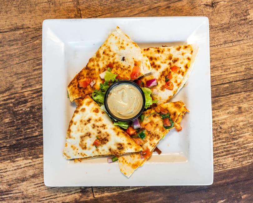 Four quesadilla wedges with pico de gallo, lettuce, and a side of dipping sauce on a white plate.