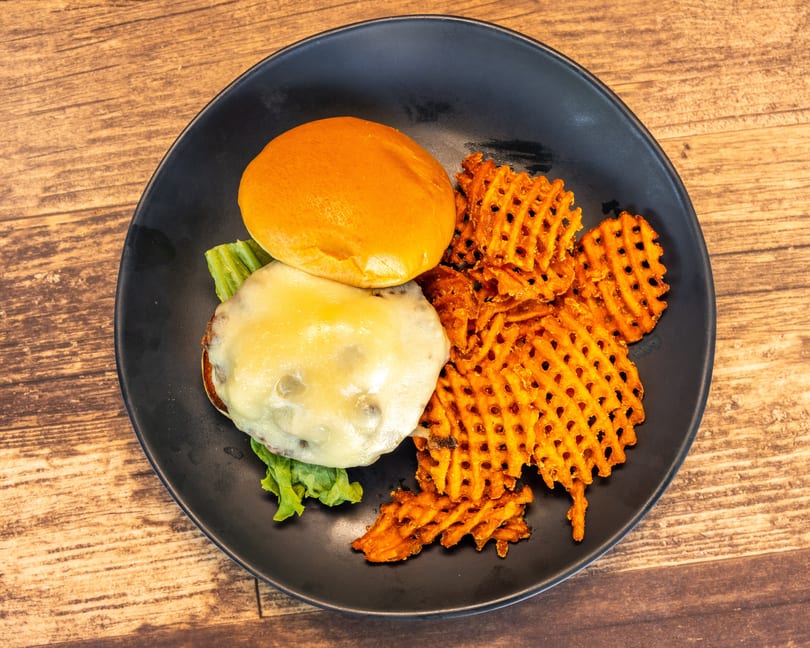Cheeseburger with melted cheese and lettuce on a bun, served with waffle fries on a black plate.