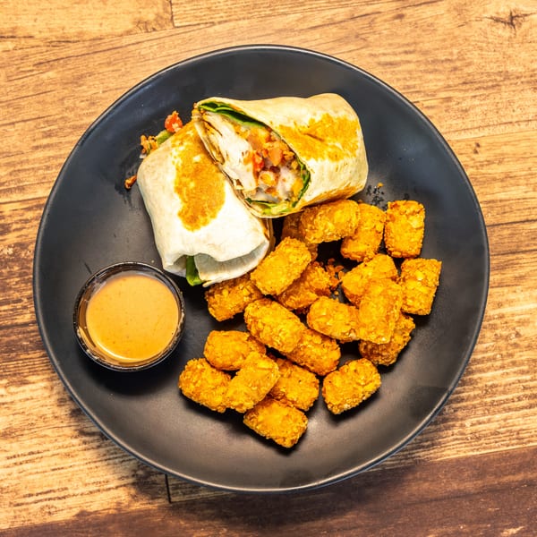 A chicken wrap cut in half, tater tots, and a side of sauce on a black plate.