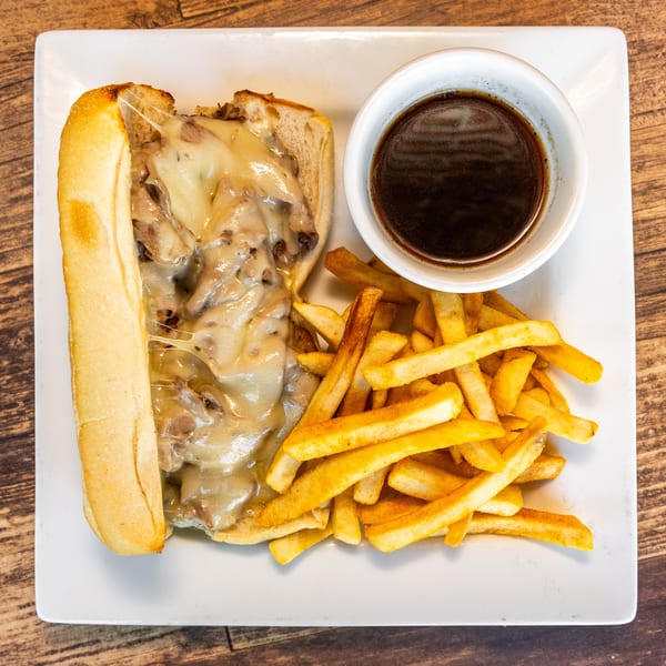 Cheesy Philly cheesesteak sandwich with a side of golden fries and a cup of au jus.