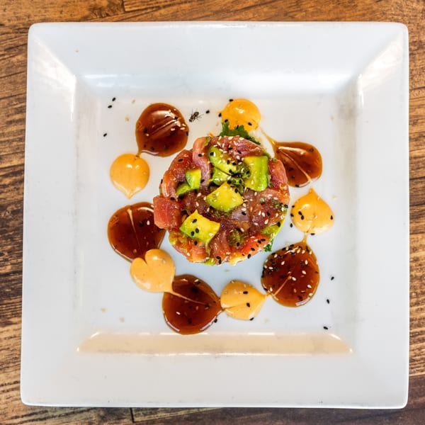 Tuna and avocado tartare with sesame seeds and two sauces on a white square plate.