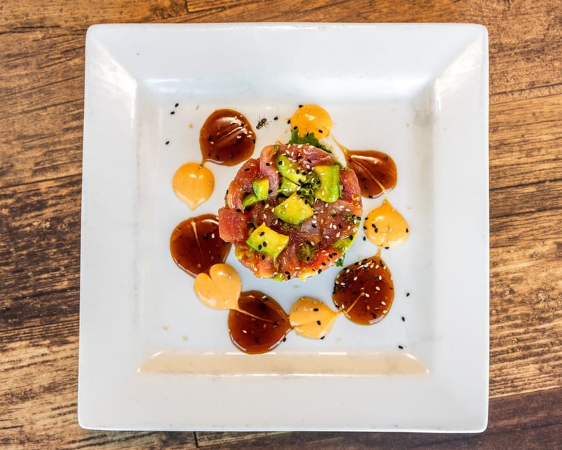 Tuna and avocado tartare with sesame seeds and two sauces on a white square plate.