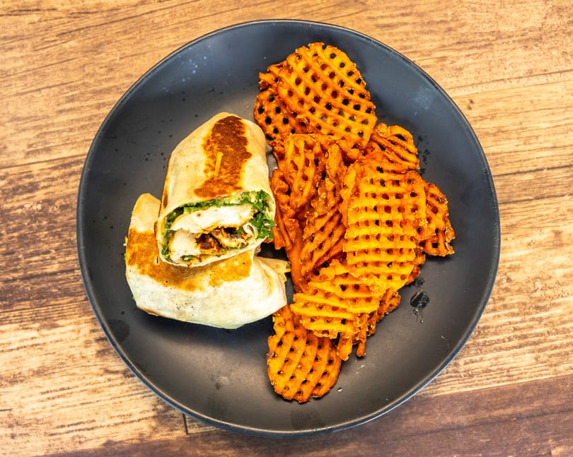 A chicken wrap with green filling and a side of waffle fries on a black plate.