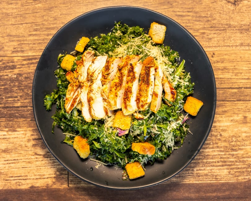 Grilled chicken Caesar salad with kale, croutons, and shredded cheese on a black plate.