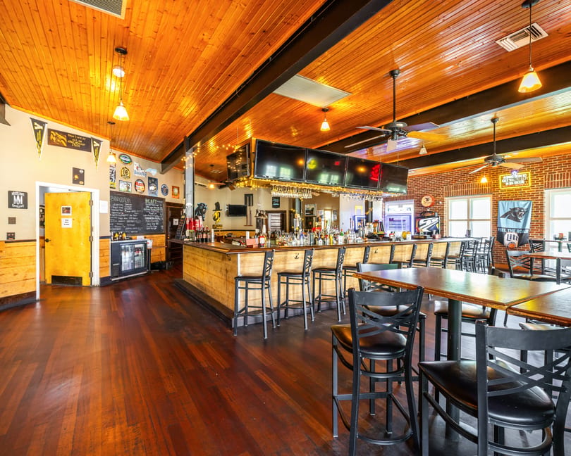 A rustic bar interior with a wooden bar, high-top tables, and a wooden plank ceiling.