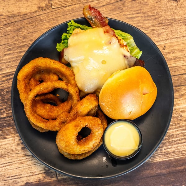 Chicken sandwich with melted cheese, bacon, lettuce, a bun, onion rings, and dipping sauce.