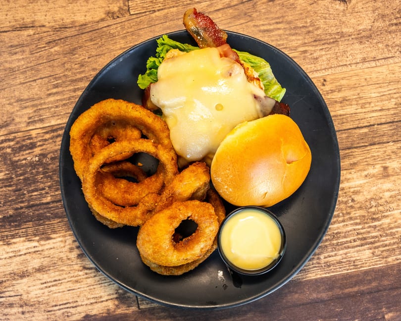Chicken sandwich with melted cheese, bacon, lettuce, a bun, onion rings, and dipping sauce.