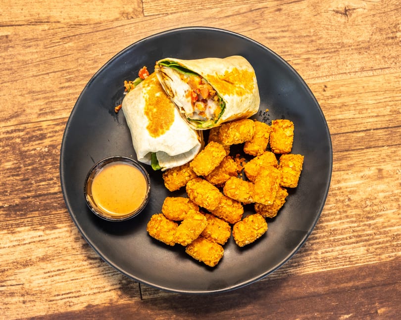 A chicken wrap cut in half, tater tots, and a side of dipping sauce on a black plate.